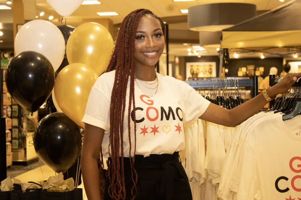 Raven Smith stands in front of a t-shirt rack. She is smiling and wearing a shirt that reads, “Go COMO.”