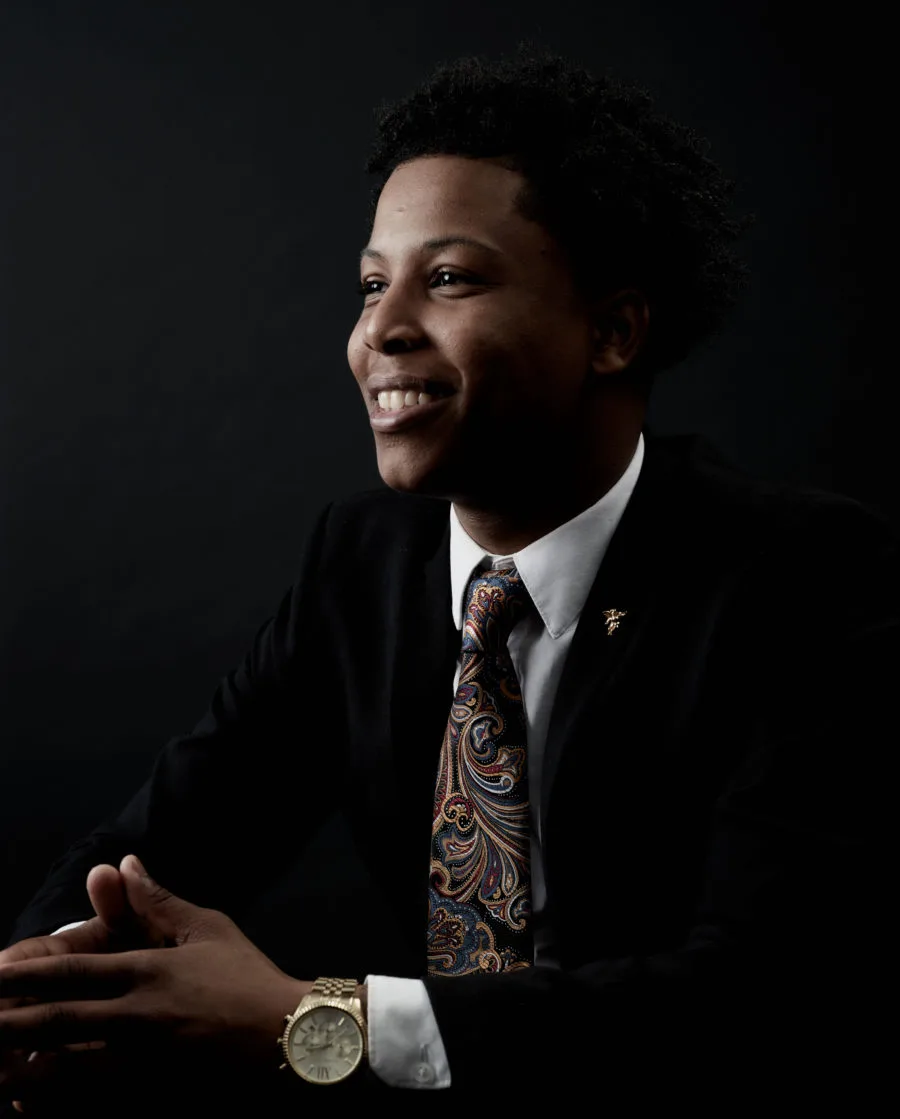 A portrait photo of a young man with a medium-deep skin tone wearing a white button-up shirt, black blazer, and multi-color, patterned tie smiles toward the left-hand side of the photo in front of a gradient black background. 