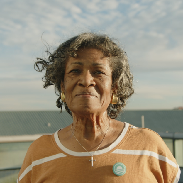 The image is a gif of Willa Marie Fox Burton smiling. Willa has a medium deep skin tone and gray hair curled to her shoulders. She is wearing gold hoop earrings, a silver cross necklace, and an orange and white striped t-shirt.  On the right side of her shirt is an Obama Foundation "Rising Sun" logo pin in the color blue. She is standing outside in the daytime with the Chicago skyline in the background.