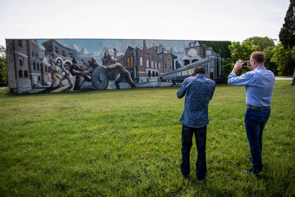 Two men standing in a plain grass field take a picture with their phones of a large, long painting. The man on the left has deep skin and really short black hair while the man on the right has light skin and blonde hair. Both are wearing dress shirts and jeans however the man on the left has his shirt untucked. The painting shows men in mechanical clothing pushing a large train wheel. In the painting background are buildings and a rail train on the right. 
