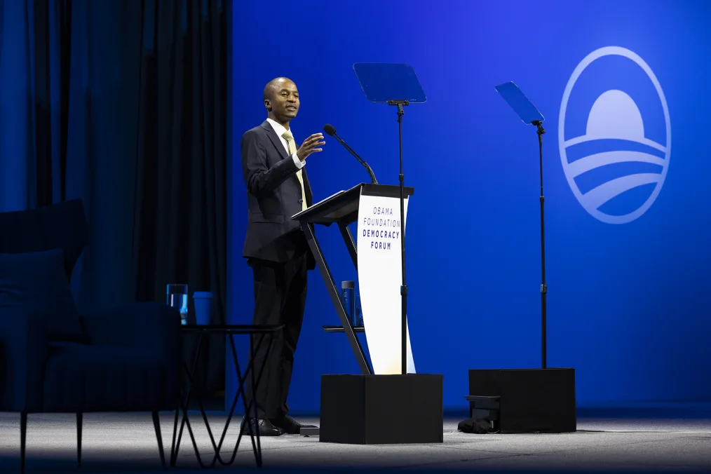 A man with deep skin stands at a podium on stage giving a speech. In the background are blue curtains and a blue wall with the Obama Foundation logo. The man is wearing a dark blue suit, white button up, yellow tie, and black shoes. Infront of the podium is a sign that reads "Obama Foundation Democracy Forum."
