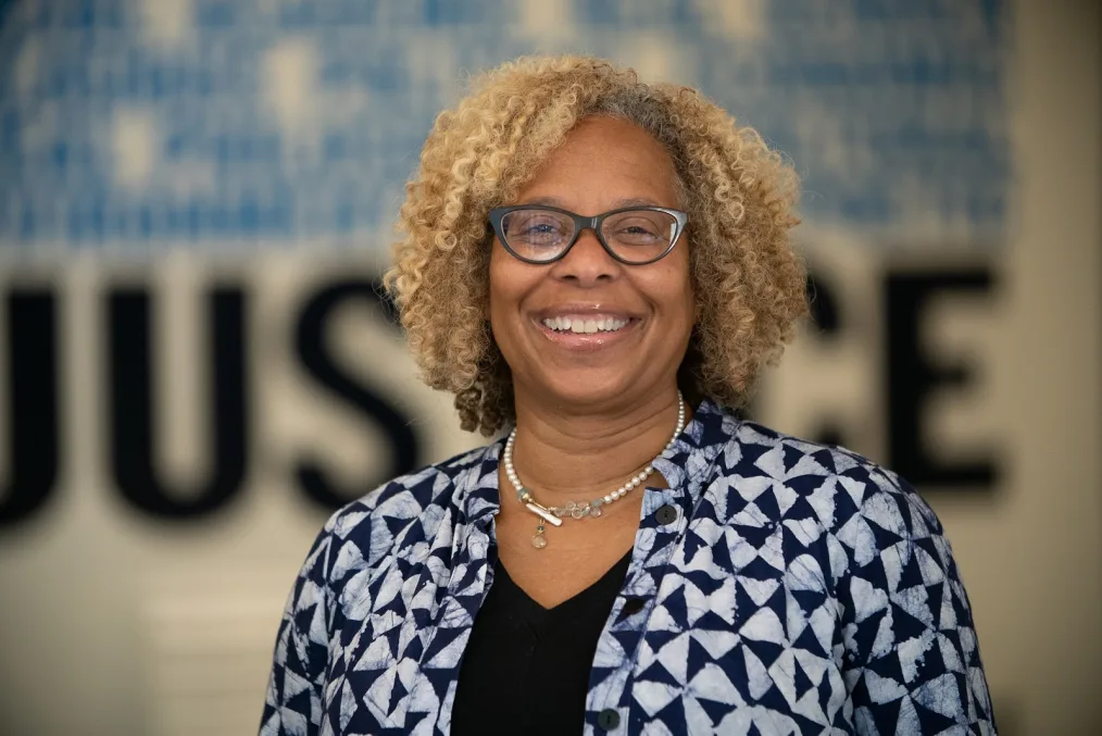 A headshot of a woman with medium-dark skin tone and blonde, curly, shoulder-length hair. She smiles broadly for the camera and wears glasses, a necklace and patterned shirt.