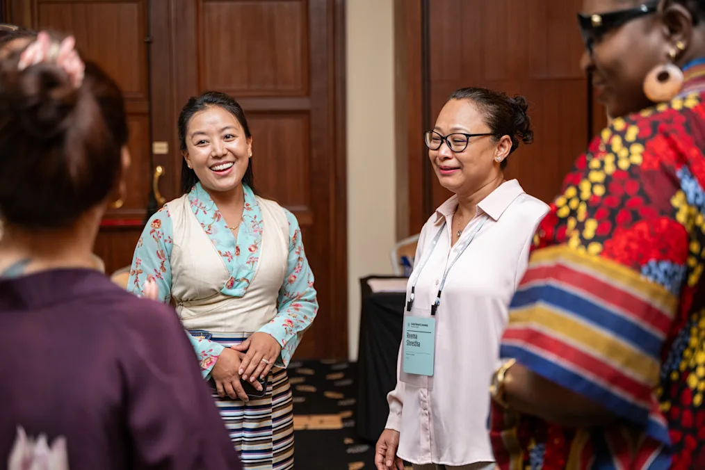A group of four women with a range of light to dark skin tones stand in a circle. All are smiling and dressed professionally.