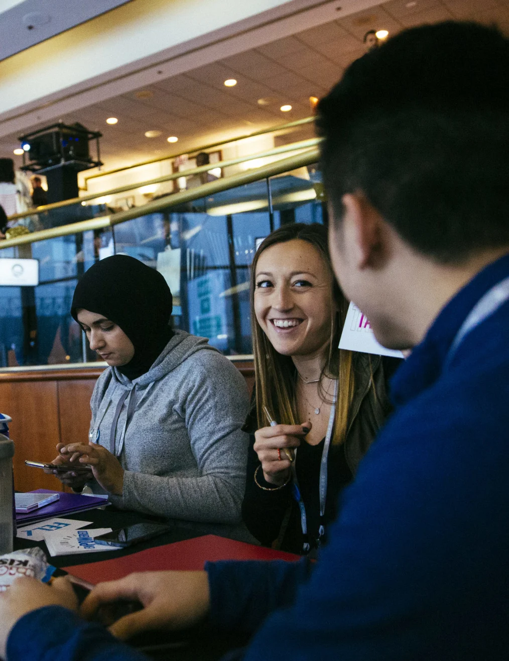 Three people with light-medium skin tones are dressed in business casual while sitting at a small table
A lady is smiling toward another light-medium skin toned man.