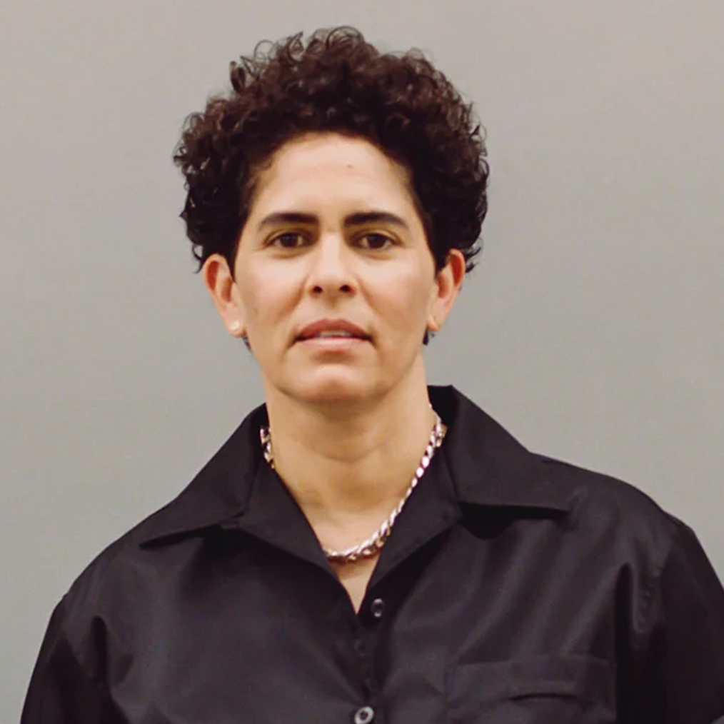 Julie Mehretu, a woman with a light skin tone and short curly black hair, holds a closed lipped smile. She is wearing a black button up shirt. 
