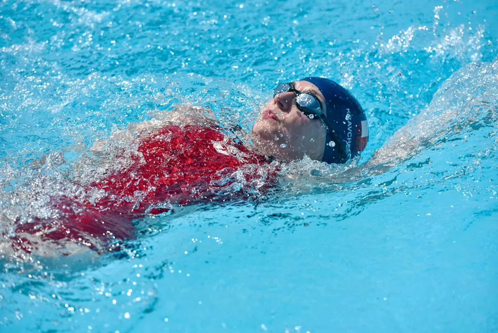 Vladyslava Kravchenko, a woman with a light skin tone, swims in a pool. She is wearing a red suit, blue swim cap, and blue goggles.