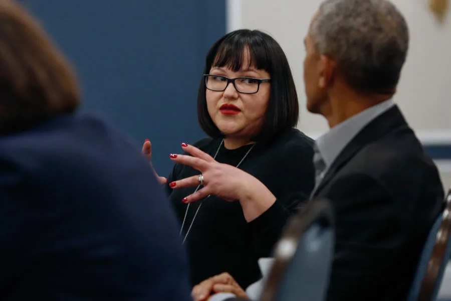 A woman with a light olive skin tone talks to President Obama. His back is to the camera. She has a short black bob, glasses, and is wearing red lipstick.