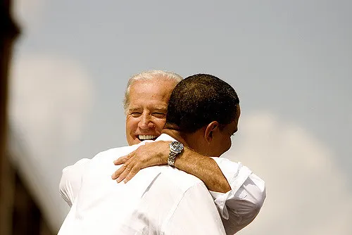 Joe Biden and Barack Obama embrace in a hug. Biden's face can be seen smiling and he wears a watch on the arm around Obama, who faces away from the camera.