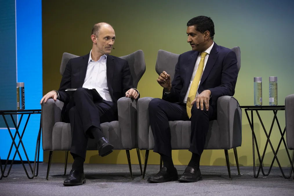 Two men sit on a grey carpet stage sitting in comfertable grey chairs. The man on the left has light skin and is bald on the top with greyish brown hair on the sides. He is wearing a suit with a white button up, black shoes, and no tie. He is engaging in a conversation with the man on the right. This man has medium-deep skin, slick black hair, a dark blue suit, white button up, spotted yellow tie, and black shoes. Coming from behind his ear is a small microphone. 