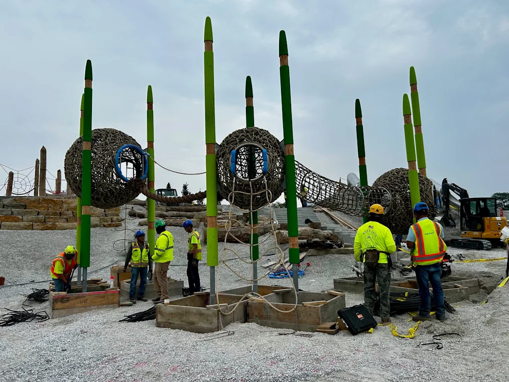 An image of construction on the Obama Presidential Center Playground. In the image, several construction workers are working on a large green playset installation. The playset installation includes several tall green pillars, netted hideaways for children to play, and a long netted "bridge" for children to walk and crawl through.