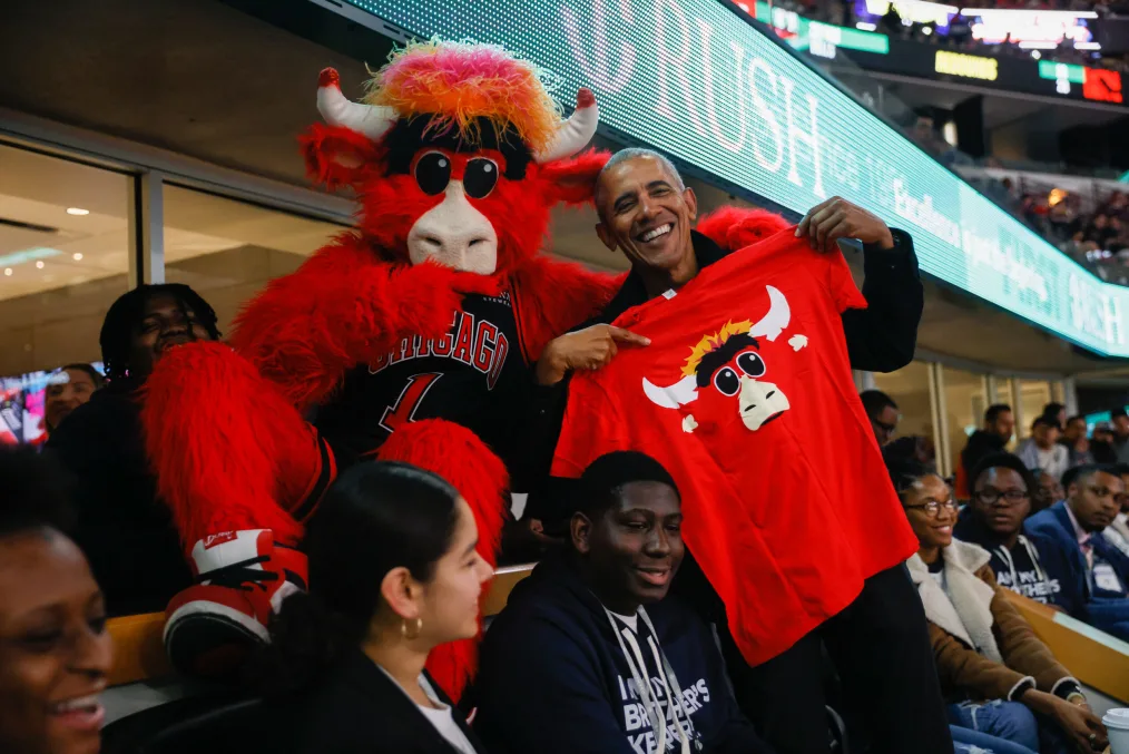 President Obama smiles, holds up and points to a red Benny the Bulls tshirt in the United Center arena. Benny the Bull mascot has one arm around President Obama.