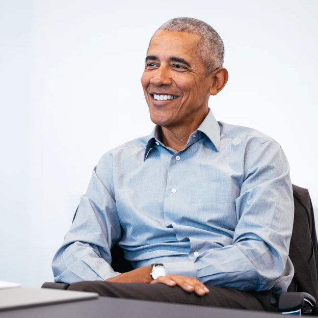 President Obama sits in a chair in a bright room and smiles off camera. He is wearing a light gray button down shirt.