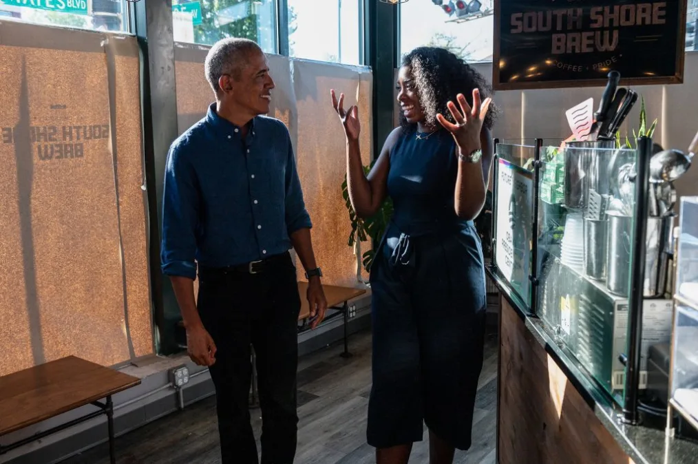 A woman holds her hands in the air, gesturing while speaking to a smiling President Obama