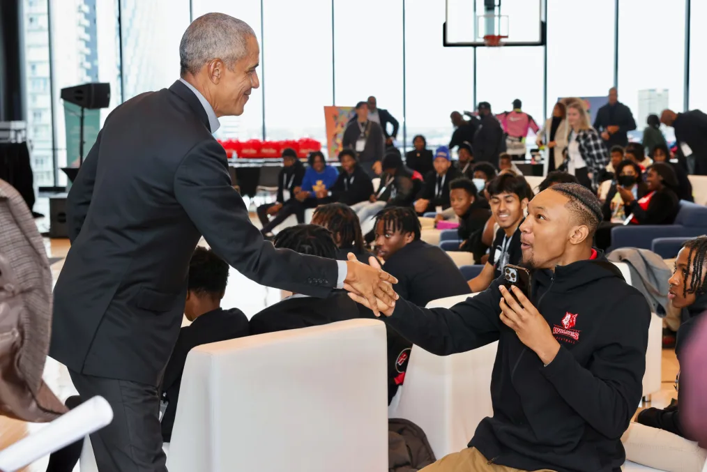 President Obama shakes hands with a seated young person with medium skin and an excited expression. A crowd of young people with various skin tones watches.