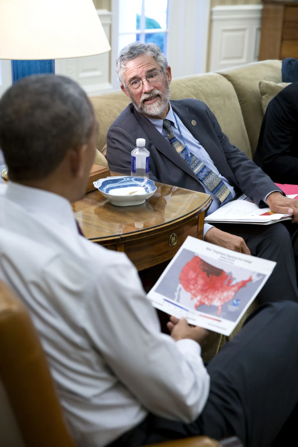 The camera focuses on an older man with a light skin tone and short, gray hair and glasses sitting on a couch in business attire. He is in the middle of a conversation with President Obama as President Obama sits in front of him holding a map in red. 