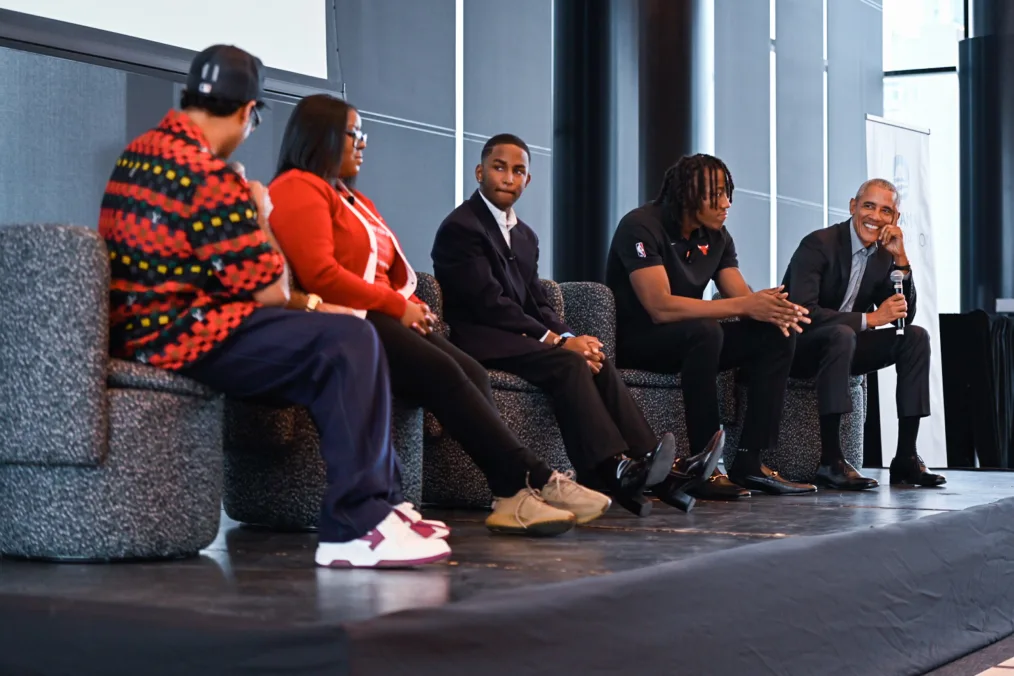 A close up photo President Obama, three men, and one woman sitting in black chairs on a small stage. The four people have deep skin complextions and are using mics to talk with each other. There attire is a mix of business causal and casual.