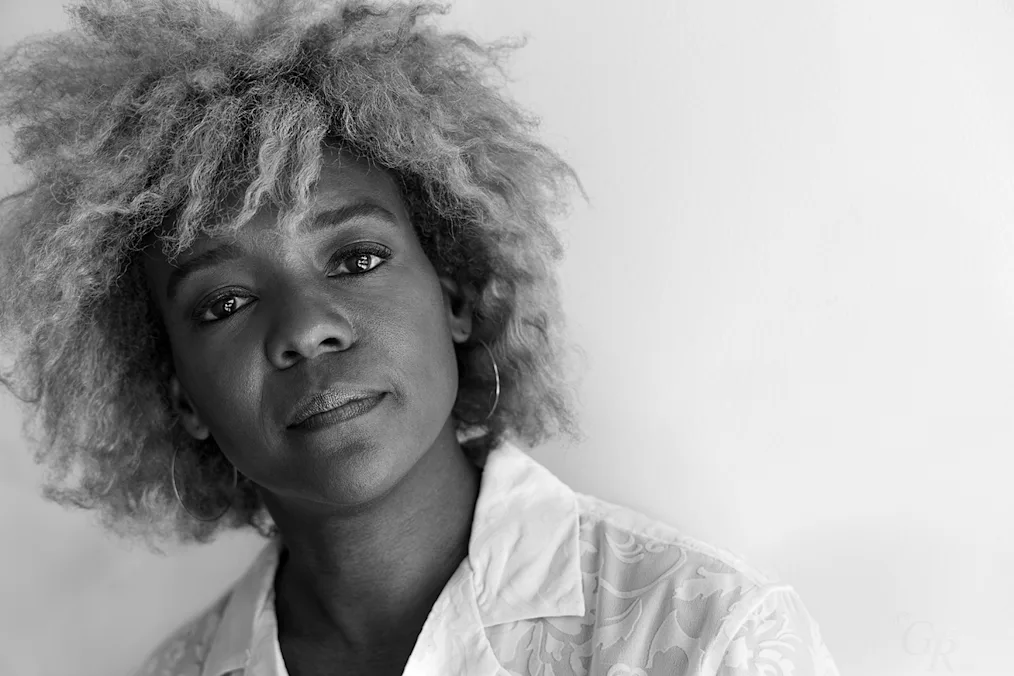 he image is a photographed portrait of Nekisha Durrett. The image is in black, white, and greyscale tones. Nekisha has a deep complexion and light colored hair that is falls in waves around the nape of her neck. She is wearing hoop earrings and a white collared shirt with a embroidered design.
