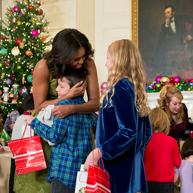 Mrs. Obama hosted the children of military families in the State Dining Room of the White House. Mrs. Obama embraces a young boy with a light skin tone at the White House. A young girl with a blue dress and blonde hair stands beside them. In the background are more children with a range of ages and light to dark skin tones. 