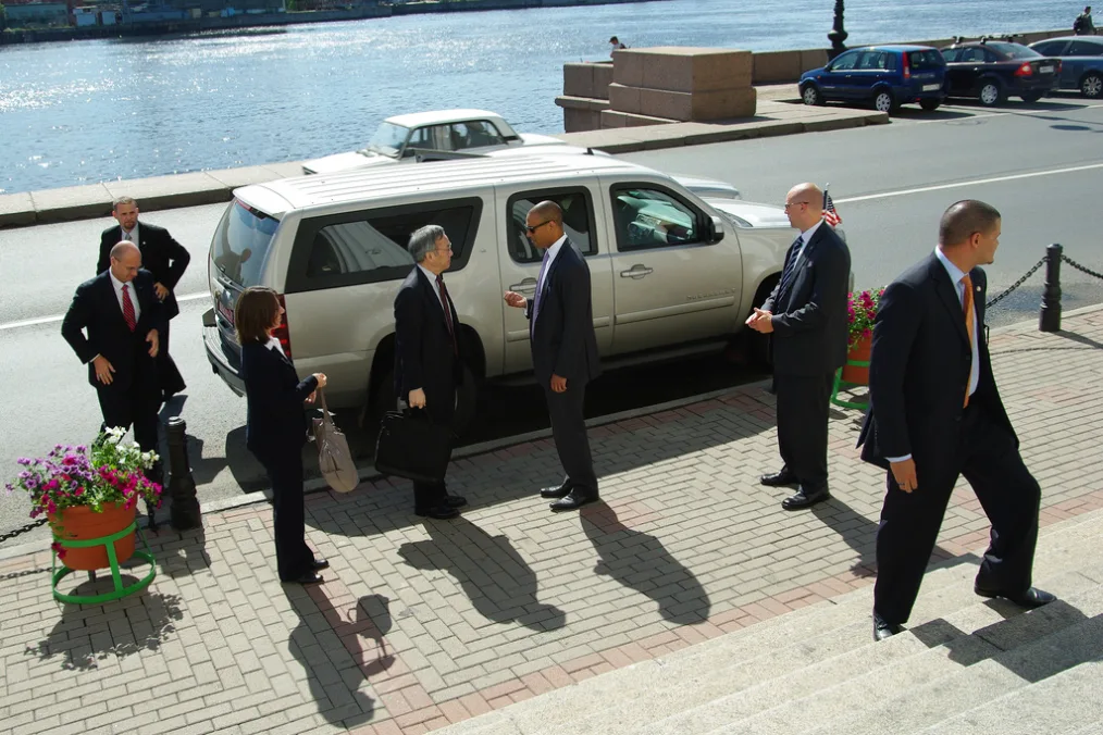 On a sunny day on a brick street along a body of water, Devin Hampton stands in front of a silver SUV and briefs Steven Chu, Former United States Secretary of Energy. Five other personnel stand around them, all wearing business suits. 