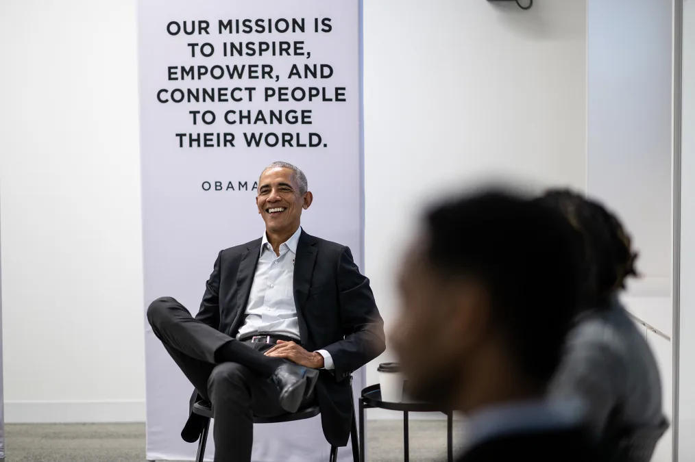 President Obama smiles as he sits with his legs crossed. A sign behind him reads, “Our mission is to inspire, empower, and connect people to change their world” and “Obama Foundation.” Out of focus to the right, two people sit side by side.