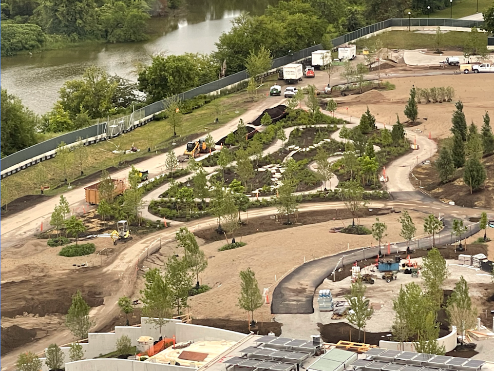 The image is an aerial view of the external Wetland Walk on the Obama Presidential Center campus. The image features several trees across the landscape and walking paths across the picture. To the left of the landscape is a body of water.