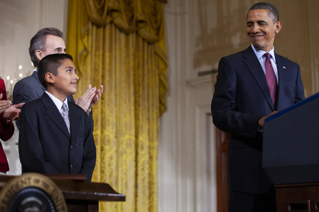  A young boy smiles at President Obama as he stands behind a podium. The boy has a medium skin tone and is wearing a suit and gray tie. Two people stand behind him in the background. 