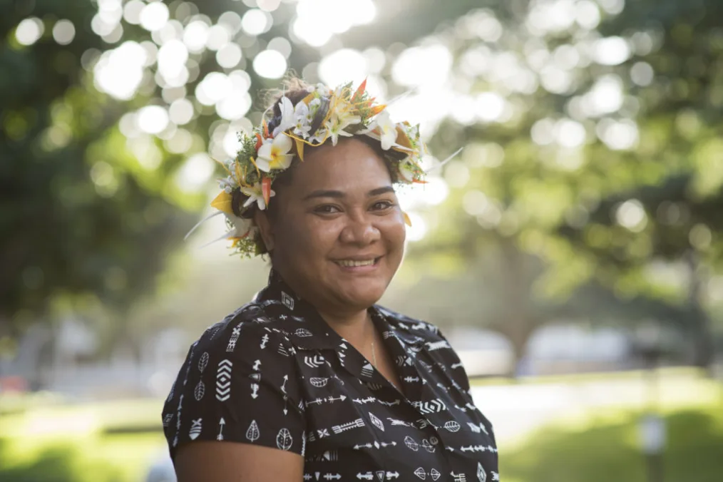In this portrait, a woman with a medium skin tone wears a flower crown lei and a dark patterned shirt smiles toward the camera.