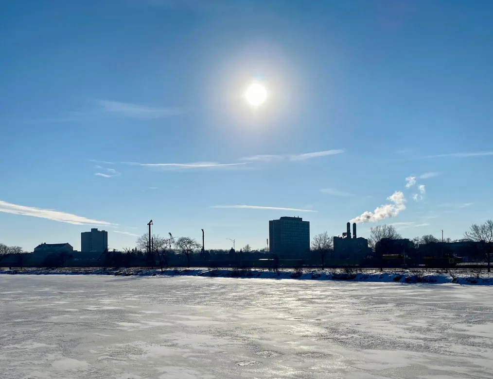 A wide shot of snow-covered ground and buildings in the background. The sun is shining in a blue sky.