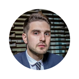 Alex Soros looks to camera. He has a light skin tone and medium brown hair. He is wearing a blue suit and navy striped tie.