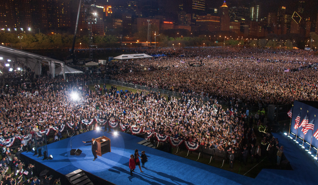 A huge crowd stretches across a large park, lit up by bright lights. At the lower left-hand corner, President Barack Obama waves at the crowd, while his wife and daughters walk off a large stage carpeted in blue. 