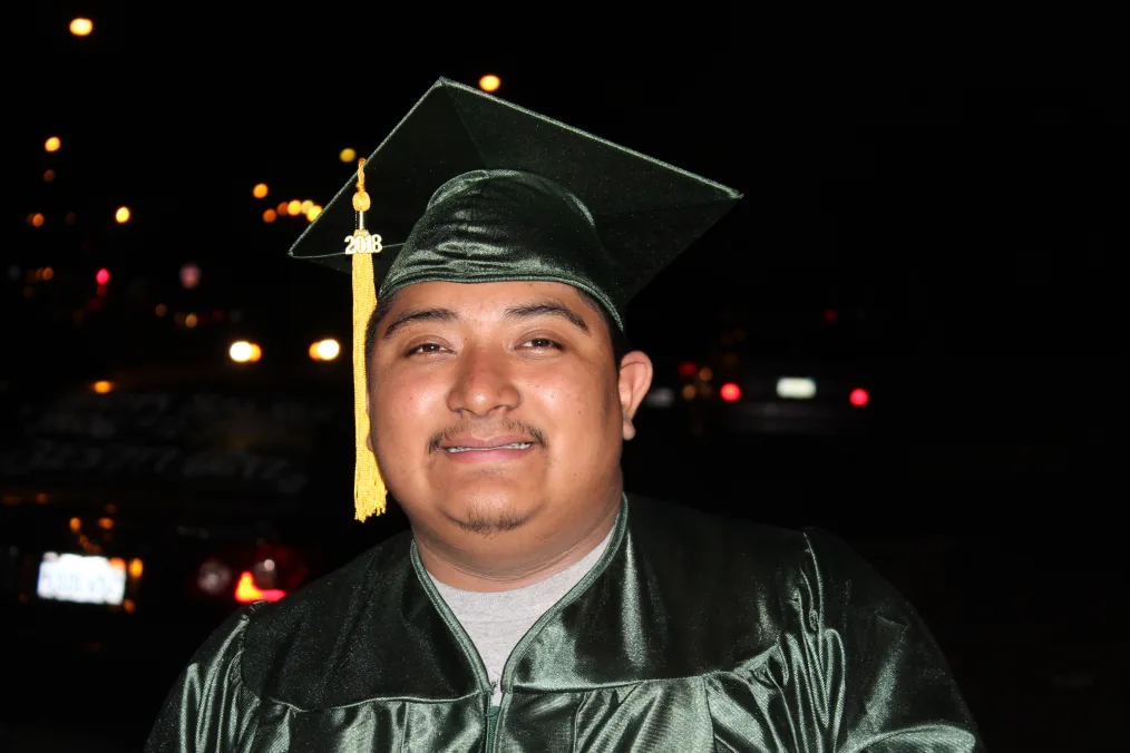A man with a medium skin tone stands smiling at the camera in a green graduation cap and grown