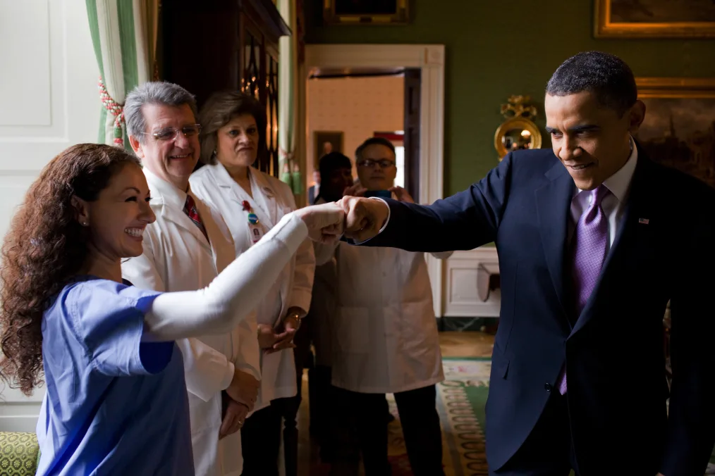President Obama fist-bumps a medical professional in the Green Room of the White House. The woman President Obama fist-bumps has a medium skin tone and long curly hair. She is wearing blue scrubs. Next to her, three doctors with a range of light to medium skin tones, smile as they observe.