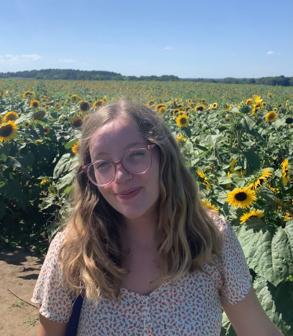 On a sunny day, Leah Whitmoyer, a woman with a light skin tone smiles as she stands outdoors. She has long blonde hair and is wearing glasses. A field of sunflowers is in the background. 