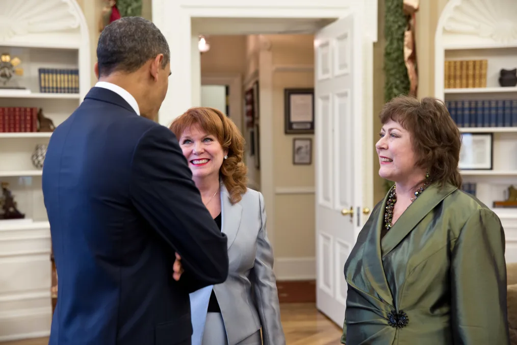 President Barack Obama talks with Natoma Canfield, a woman with a light skin tone and short brown hair, and her sister, Connie Anderson, a woman with a light skin tone and long red hair, in the Oval Office. All are wearing suits.