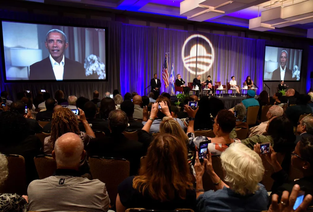 A room full of various people sit in chairs as they record and take pictures of video playing a large screen of President Obama as people sit on a panel in the far back as the Obama Foundation logo is projected 