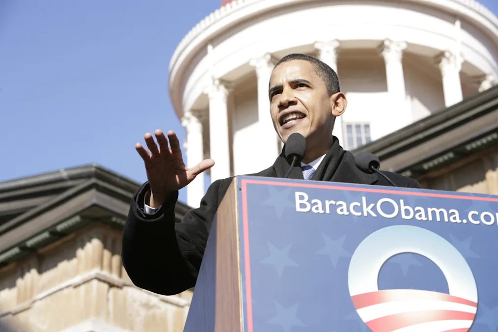 President Obama extends his hand as he stands behind a podium. On the podium is a sign that reads, “BarackObama.com” and features the Obama rising sun logo. The Illinois State Capitol building is in the background.