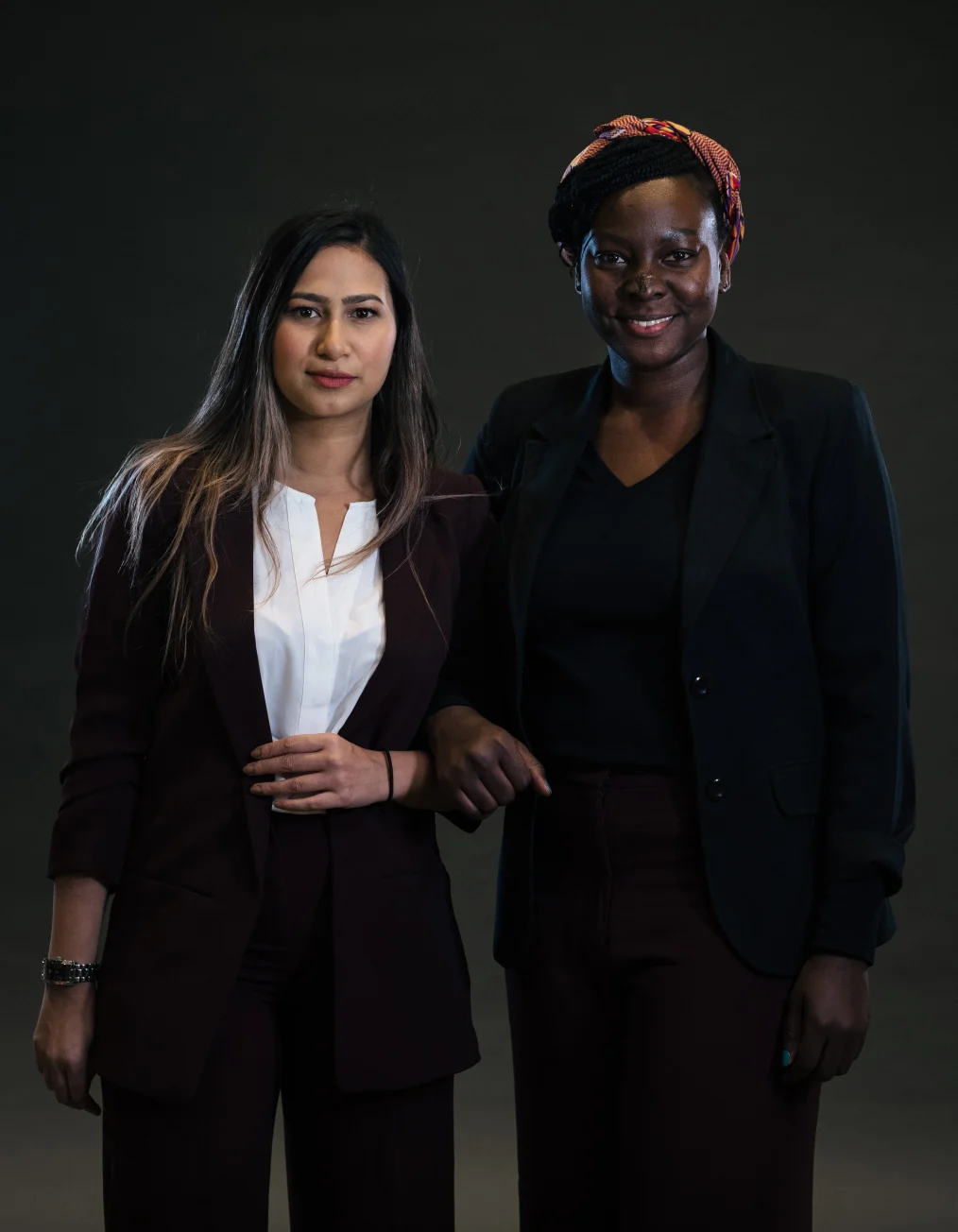 This picture shows two women with different skin tones dressed in business casual posing toward
the camera. One lady has a light-medium skin tone and wears a white shirt underneath a burgundy
suit and pants. The other lady has a deep skin tone and wears a black shirt under a black suit and
burgundy pants.