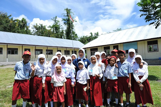 A group of young individuals with a variety of skin tones wearing white button-up shirts and red bottoms stand in the grass in front of two white buildings. 