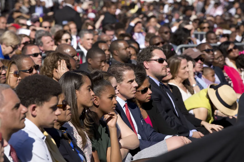 Malia Obama and Sasha Obama, a deep medium skin tone young girl, sit in a large crowd outdoors with a diverse group of people ranging from young to old and light to deep skin tones 