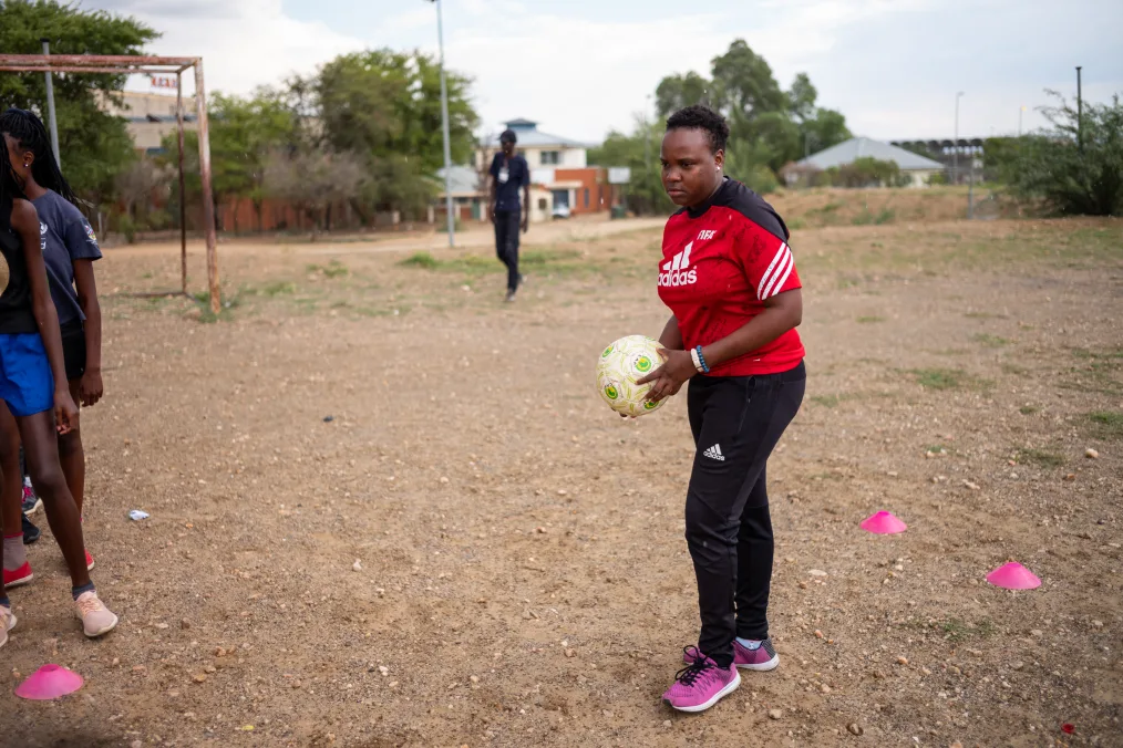 A young fairly sized women with deep skin, a small high top curly fro, and a small earing on her left ear holds a soccer ball. She is wearing a red addidas shirt, black adidas pants, and pick purple shoes. The setting is a rocky dirt field with short purple cones on the ground. A few deeper toned young girls with young hair to the side cut out by the frame. There is a rusted goal also cut out on the same side behind them. 