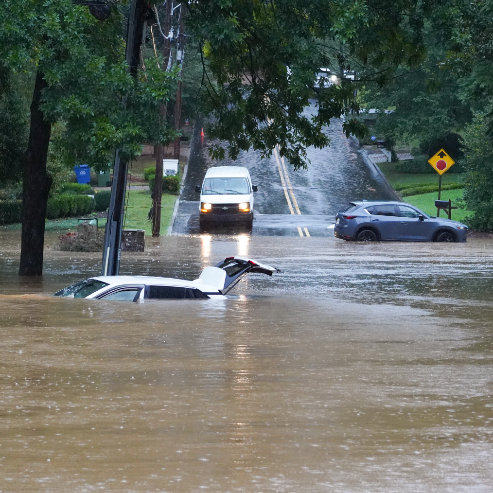 The image is a photo of a flooded street near Atlanta, Georgia due to Hurricane Helene. In the front of the photo dirty water has risen nearly above the hood of a white SUV. The white SUV has an open trunk. In the background a white van is driving down the wet and flooded street. To the right of the white van is a blue sedan car, turned horizontally, presumably stuck on the street. On both sides of the street are tall green trees.