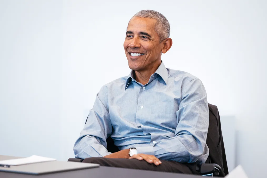 President Obama sits in a chair in a bright room and smiles off camera. He is wearing a light gray button down shirt.