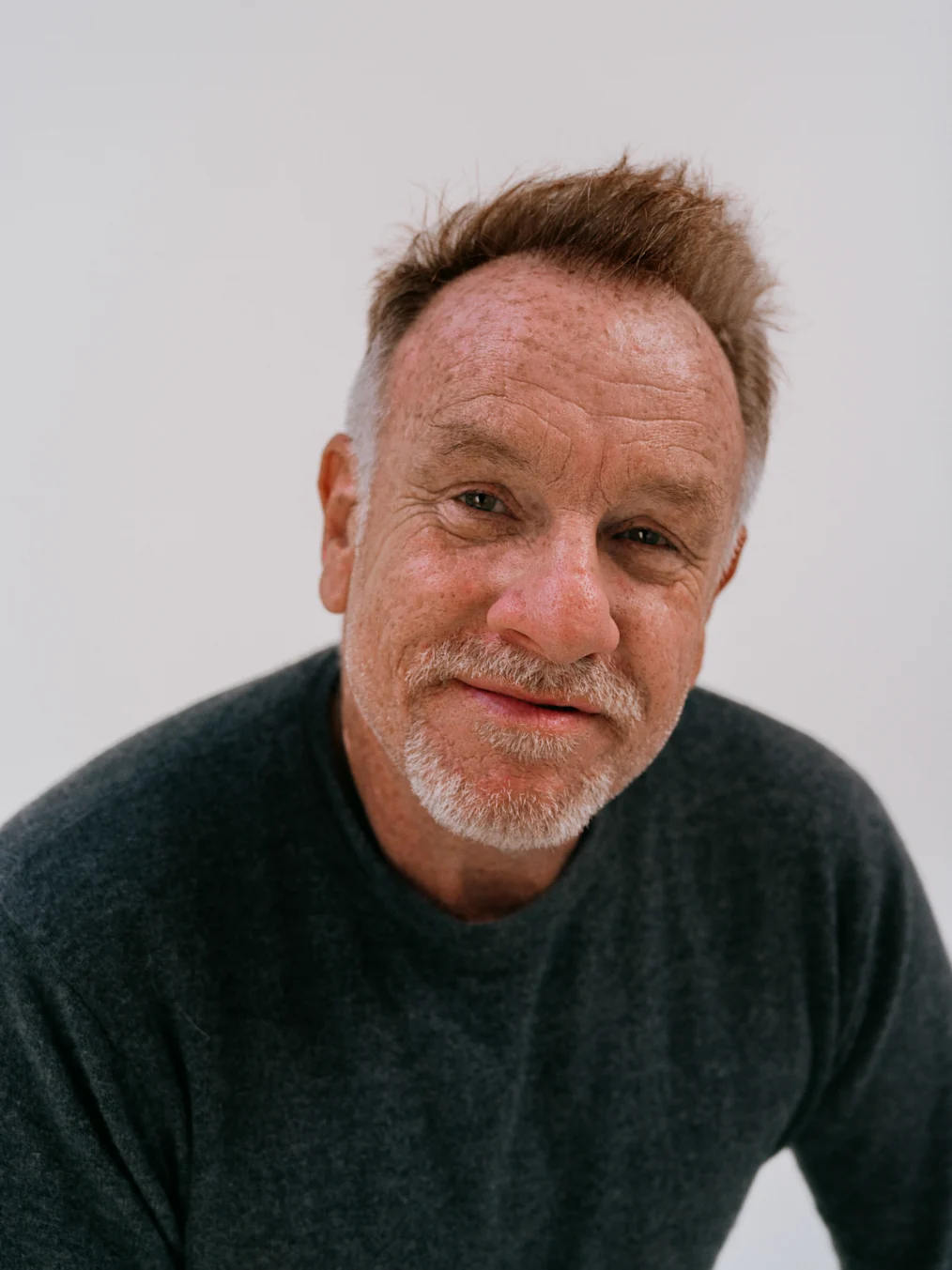 The image is a photographed portrait of Jack Pierson. Behind Jack is a white wall. Jack is staring into the camera. He has an olive complexion, short light colored hair, and a goatee. He is wearing a dark colored sweater.
