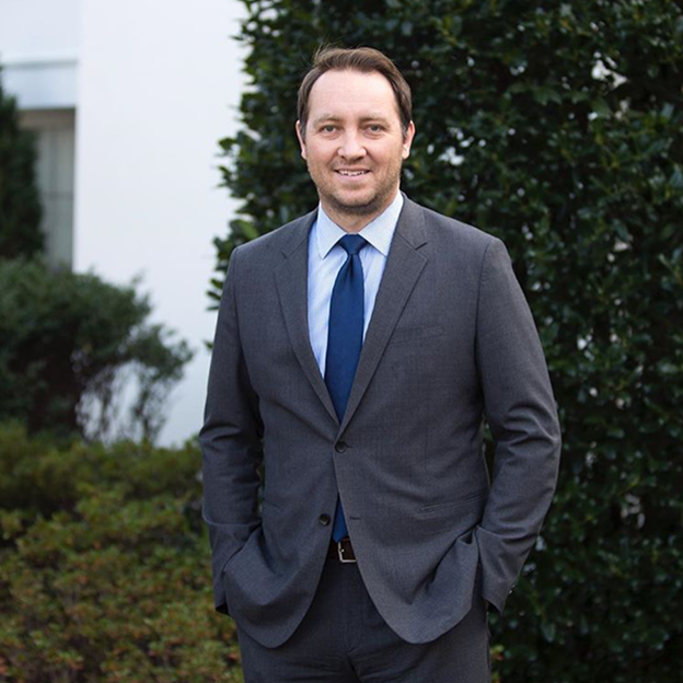 Robert Diamond, a man with a light skin tone smiles at the camera. He is wearing a suit and is standing at the White House. 