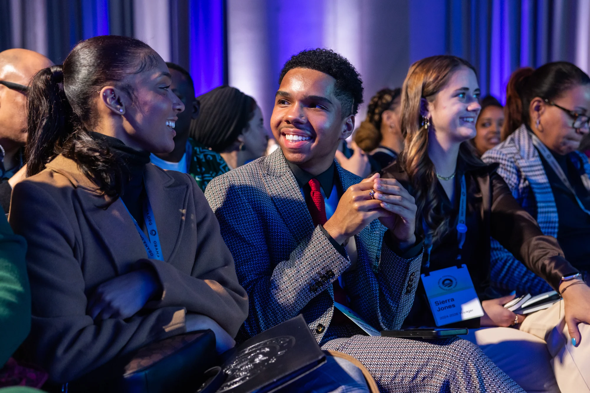 A Black male with a medium skin tone smiles at a Black woman with a dark skin tone as they sit in the audience at the 2024 Democracy Forum. A blue and white light is in the background. He is sitting around a group of people with a range of light to dark skin tones. 