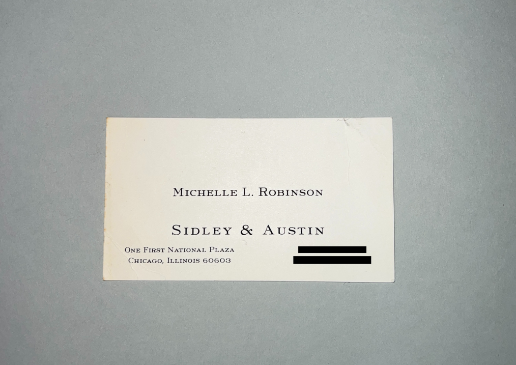 An off-white rectangular card sitting on a gray table. The off-white card is older and browning across the left side. The card is a traditional business card. On it is black text that reads, “Michelle L. Robinson, Sidley & Austin, One First National Plaza, Chicago, Illinois 60603” On the right hand side of the card, two small black boxes cover up a phone number and a fax number.
