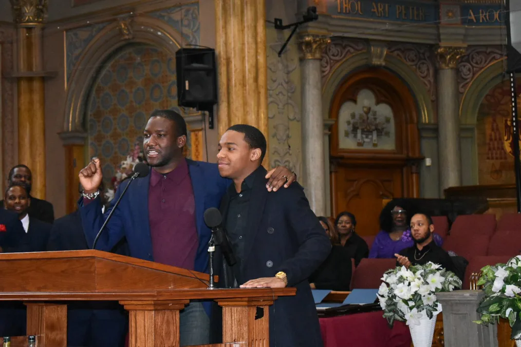 A picture of a middle aged man with deep skin tone, short hair, low chin beard, a navy blue suit, and burgandy polo underneath having is left arm around a young man with medium skin tones short hair, dark blue suit jacket, and black button up. The older man is saying something important into a mic extending from a brown stand. People sitting and standing in the background are smiling and clapping. The setting is an older church.