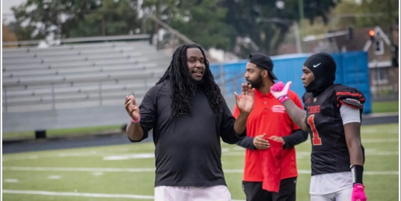 Lazarus Daniels, a Black man with a dark skin tone and long locs, is wearing a black long-sleeve shirt and white pants. He is standing on a green athletic field with his hands gesturing outward as he speaks to two other people. One of the people he is speaking to is wearing a red shirt, and the other is wearing a black football jersey with the word "LIONS" visible on the chest and the number one. Both have dark skin tones.