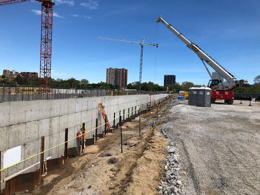 A construction site with several large cranes over a concrete wall and dirt field.