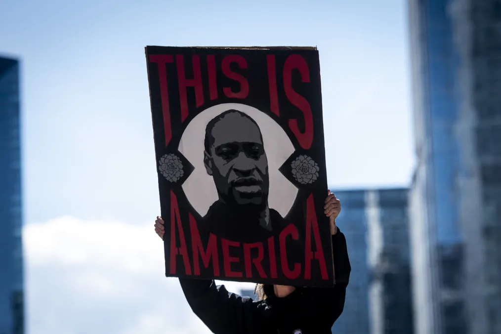 A protestor holds up a sign of George Floyd's face surrounded by the words "This is America."